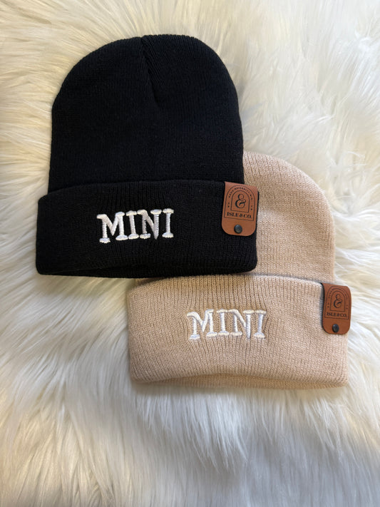Two beanies, one black and one beige, with 'MINI' branding on a fluffy white surface.