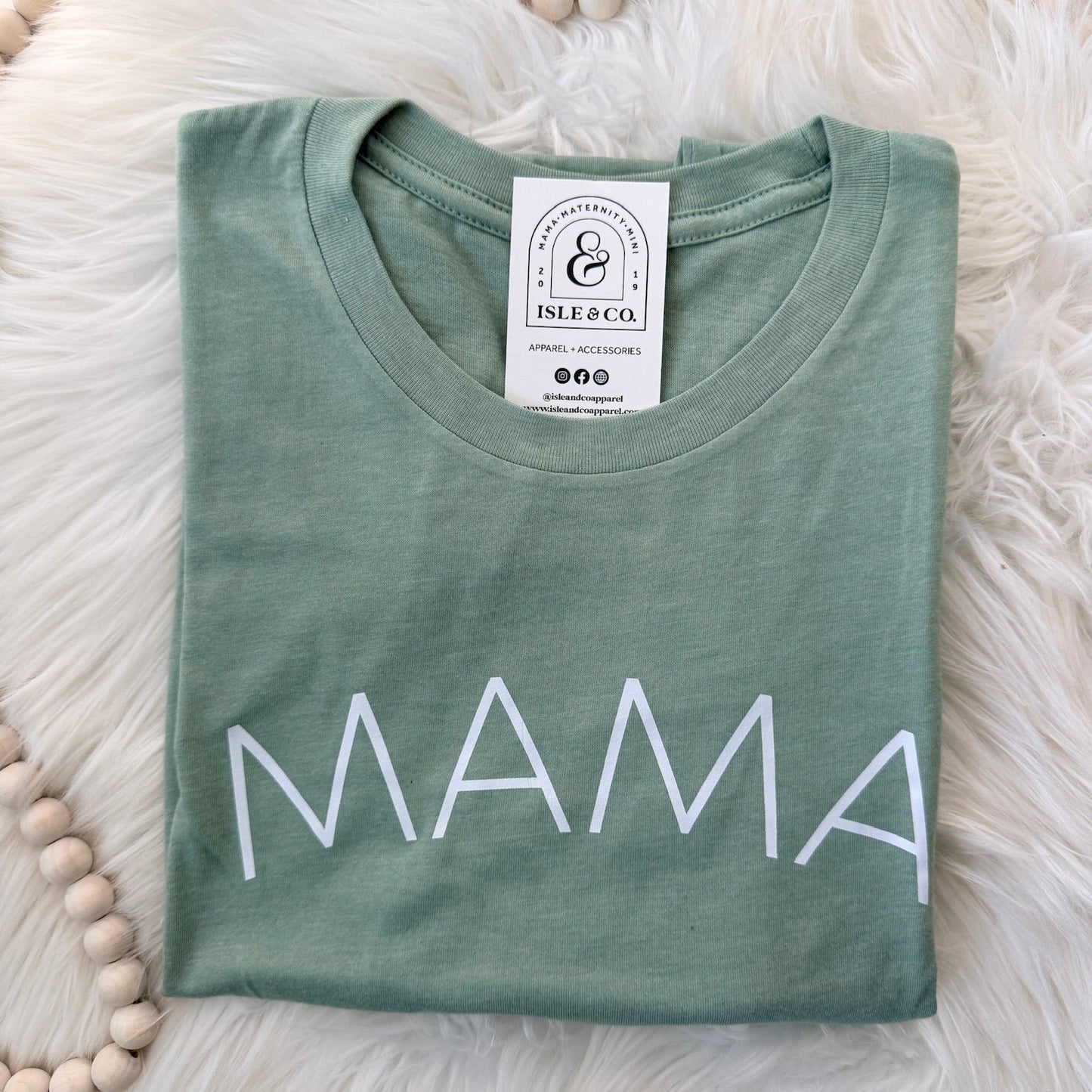 Green t-shirt with 'MAMA' printed on it, lying flat on a fluffy white surface.