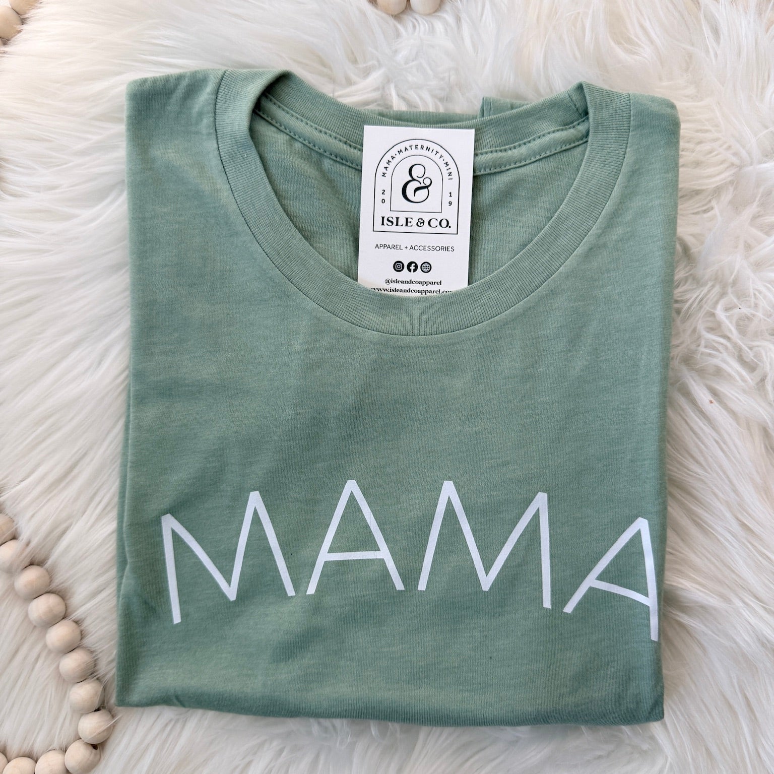 Green t-shirt with 'MAMA' printed on it, lying flat on a fluffy white surface.