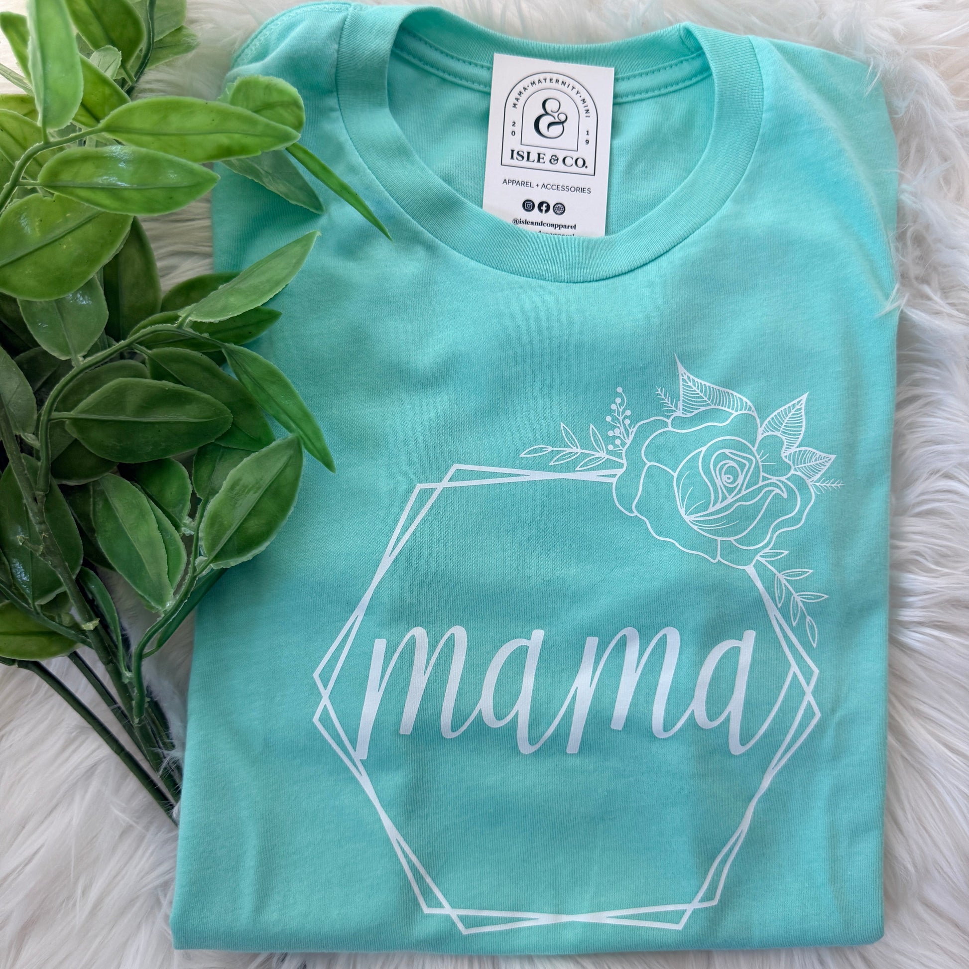 Turquoise t-shirt with white floral design and text on a fluffy white surface with green leaves.